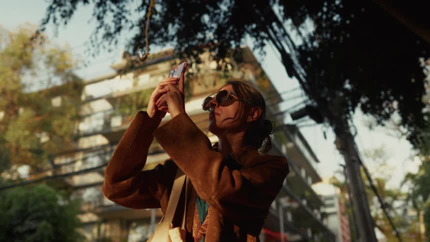 A young woman wearing sunglasses captures a photo with her smartphone on a sunny city street