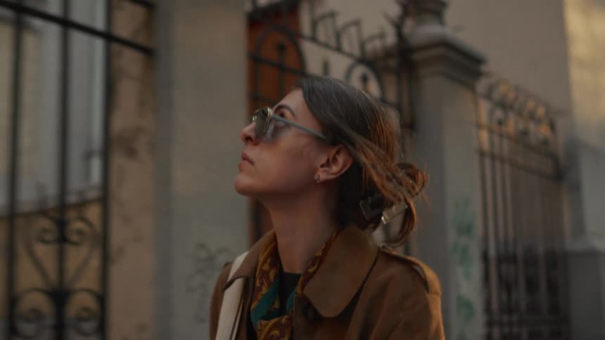 A stylish woman in sunglasses walks thoughtfully down a city street in slow motion, the camera pulling back from a close-up