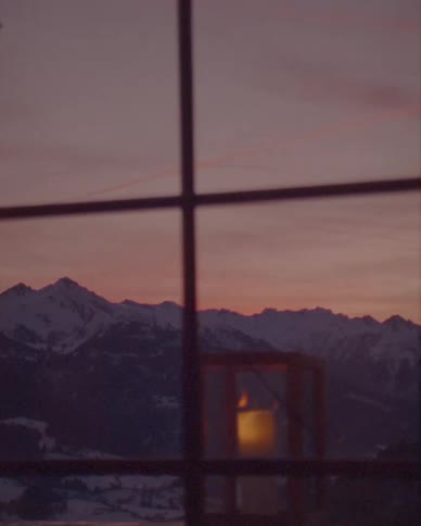 A serene window view of a snowy mountain range at sunset with a warm candle flickering in a lantern
