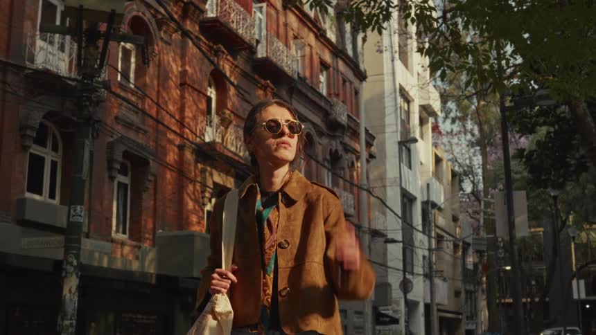 A fashionable young woman in sunglasses stops to look around while walking down a sunlit city street with beautiful architecture