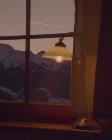 A tranquil view from a cozy cabin window shows a snowy mountain range as a warm lamp glows on the windowsill