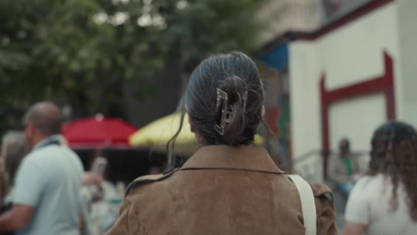 Over-the-shoulder shot of a woman in a brown jacket walking through a bustling outdoor market in slow motion
