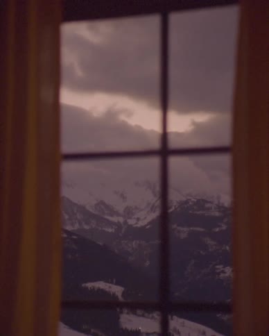 A moody, static shot of a winter mountain landscape framed by a window and curtains