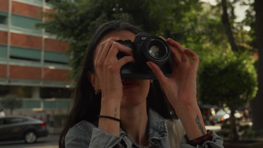 A slow push-in shot shows a young woman with tattoos focusing the lens of her camera on a city street