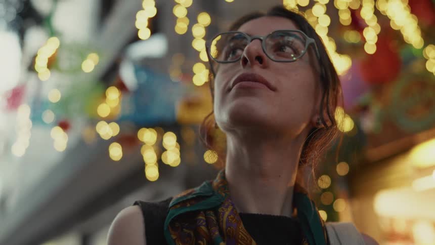 A slow-motion, low-angle shot captures a woman with glasses gazing up at festive lights and decorations on a city street