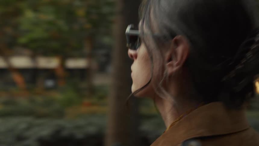 A stylish woman in sunglasses and a brown coat walks confidently through a lush city park in slow motion