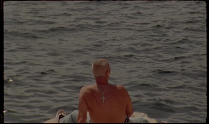 A shirtless older man wearing a cross necklace sits by the calm ocean listening to music