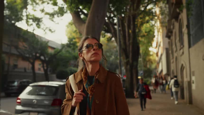 A stylish woman in sunglasses and a trench coat walks confidently down a tree-lined city sidewalk in slow motion