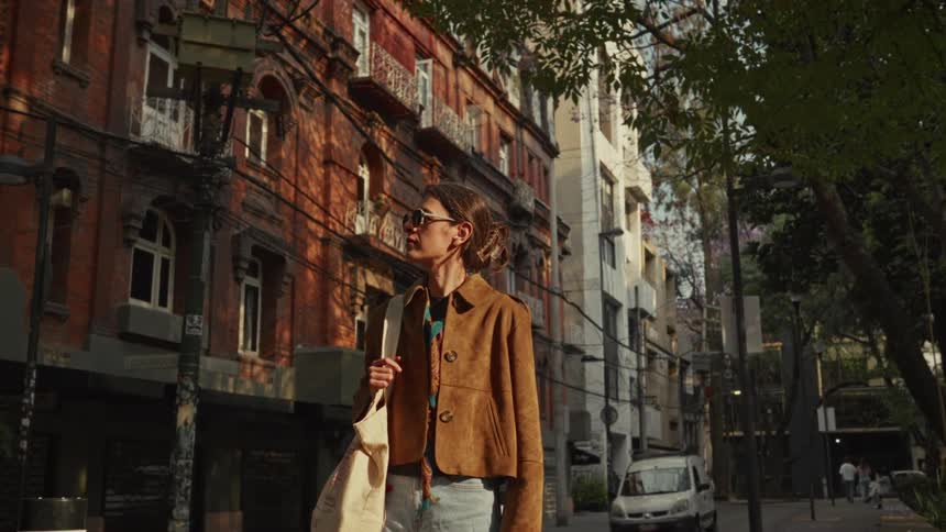 A stylish woman in sunglasses walks down a sun-drenched city street with historic brick buildings in slow motion