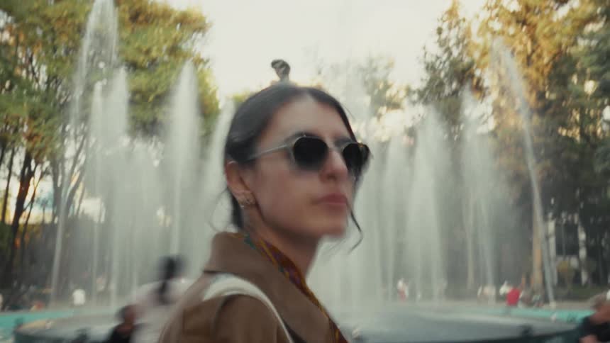 A stylish woman in sunglasses walks past a large fountain in a bustling city park on a sunny day