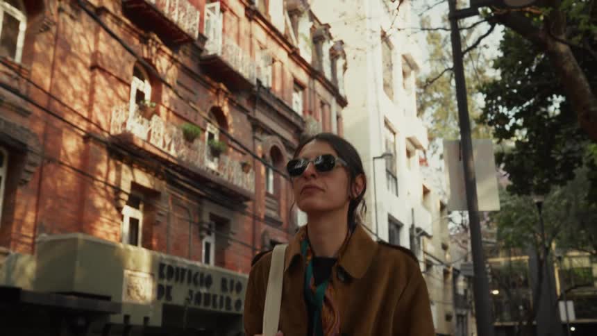 A stylish woman in sunglasses and a brown coat smiles as she walks down a sun-drenched city street in slow motion