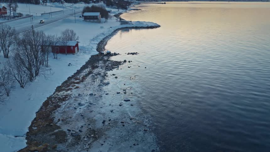 An aerial shot flies over an icy shoreline, revealing a serene, snow-covered Norwegian village and mountains at sunset