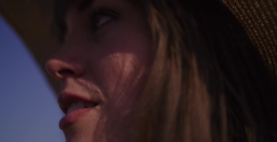 A beautiful woman in a straw hat opens her eyes in slow motion, her face illuminated by the golden hour sunlight