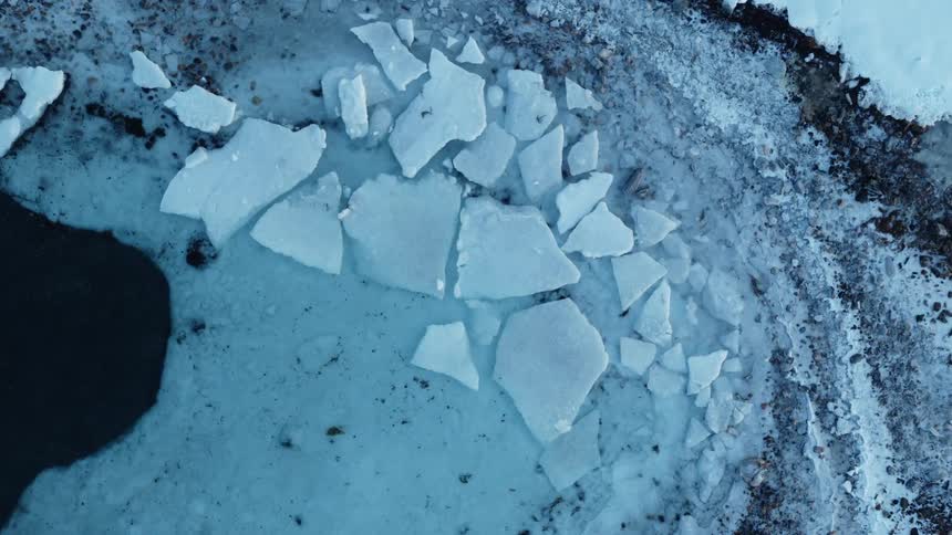 A static aerial top-down view of shattered ice floes resting on the frozen shore of a dark body of water