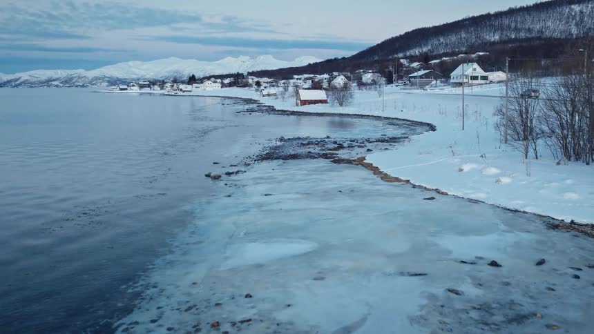 Slowly moving over the icy shoreline of a Nordic fjord, revealing a quiet, snow-covered village nestled beneath mountains at dusk