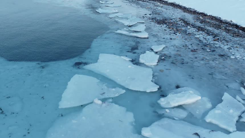 An aerial view pulls back to reveal a frozen, rocky shoreline with shattered ice sheets on the water's edge