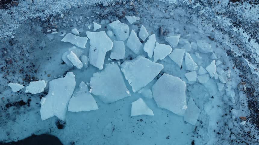 A top-down drone shot slowly ascends from a close-up of fractured blue ice to reveal a frozen, snowy shoreline