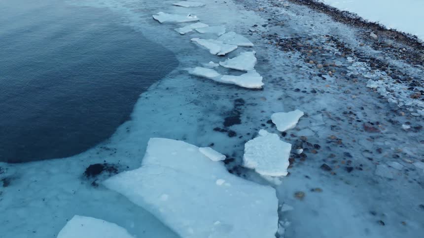 A slow aerial pull-back reveals a desolate and frozen shoreline where dark water meets cracked ice and snow-covered rocks