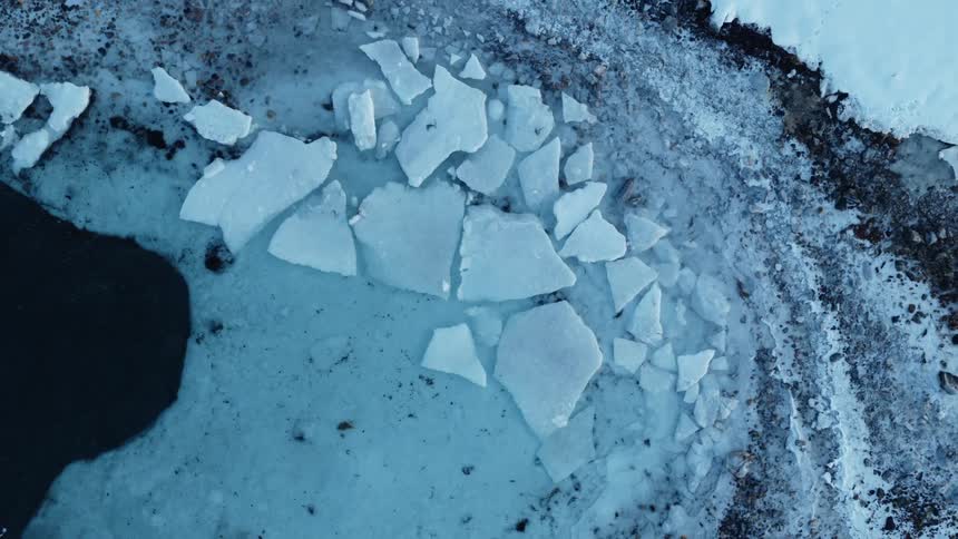 An aerial top-down shot slowly pulls out to reveal broken ice sheets along the shoreline of a frozen body of water
