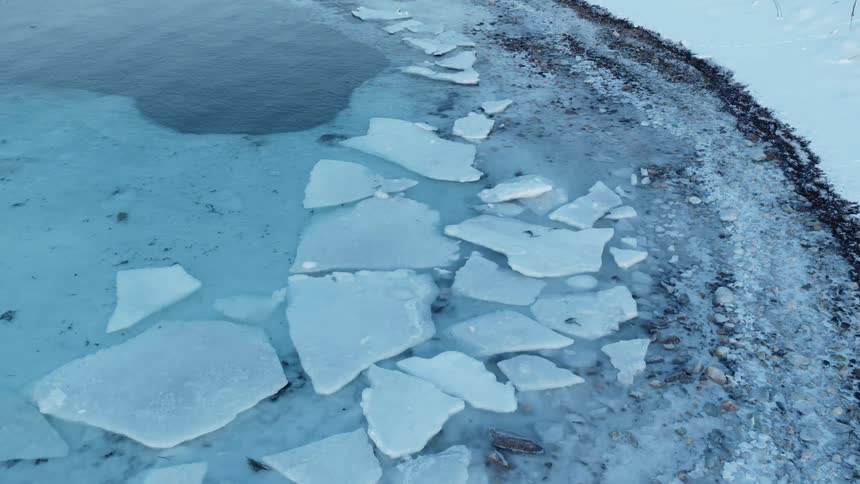 An aerial view slowly glides over the icy shoreline where broken ice sheets float in the frigid, clear water