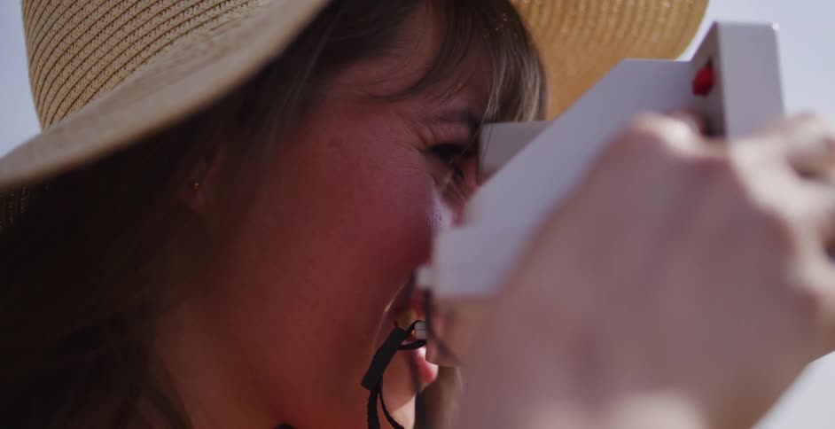 A happy woman in a straw hat smiles as she takes a picture with a vintage instant camera on a sunny day