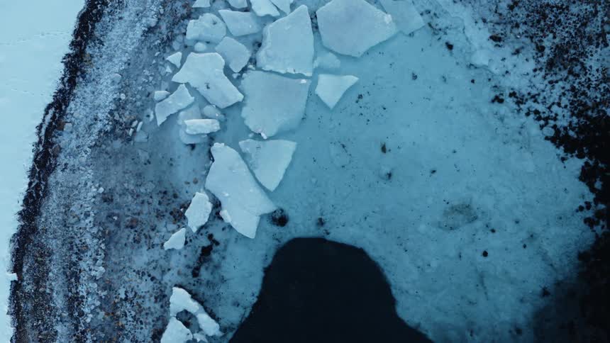 A top-down aerial view slowly pulls back to reveal a frozen shoreline with broken ice floating in the dark, cold water