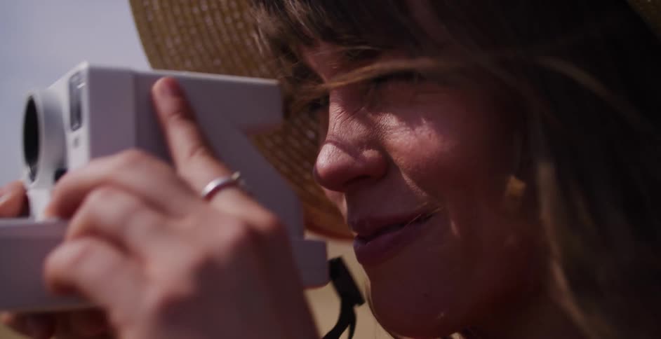 Close-up of a smiling woman in a straw hat bringing up an instant camera to take a picture on a windy day