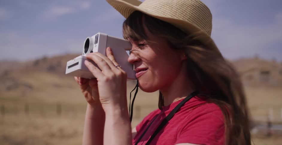 A happy woman in a straw hat smiles as she raises her instant camera to take a picture on a windy day in the country