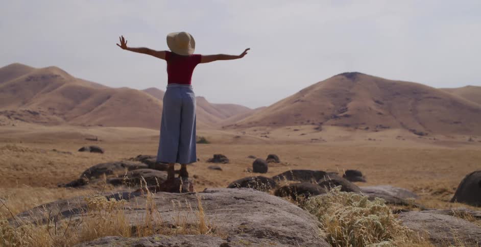 A free-spirited woman in a straw hat joyfully balances on a large rock with her arms outstretched, surrounded by a vast landscape of golden, rolling hills