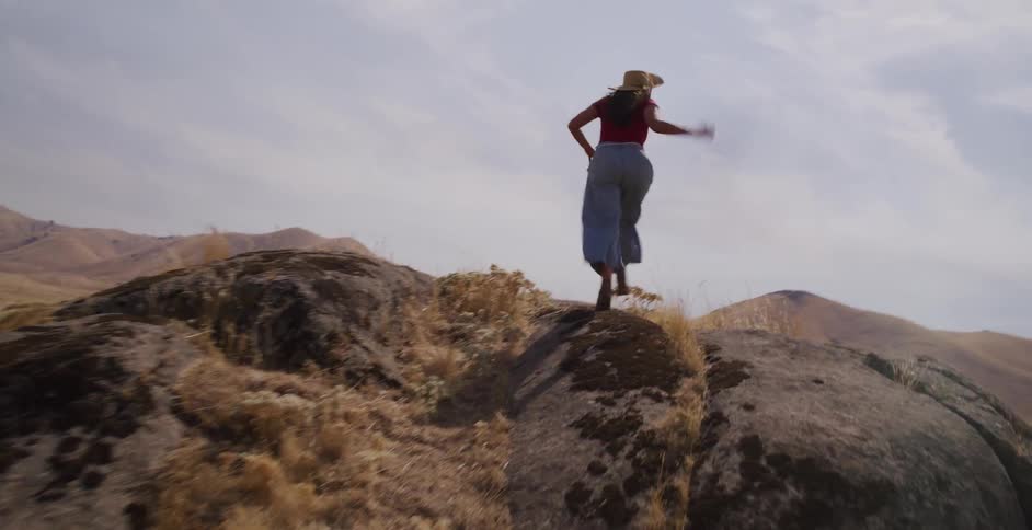 A woman in a straw hat runs up a rocky hill in a vast, arid landscape, stopping to spread her arms in a moment of freedom