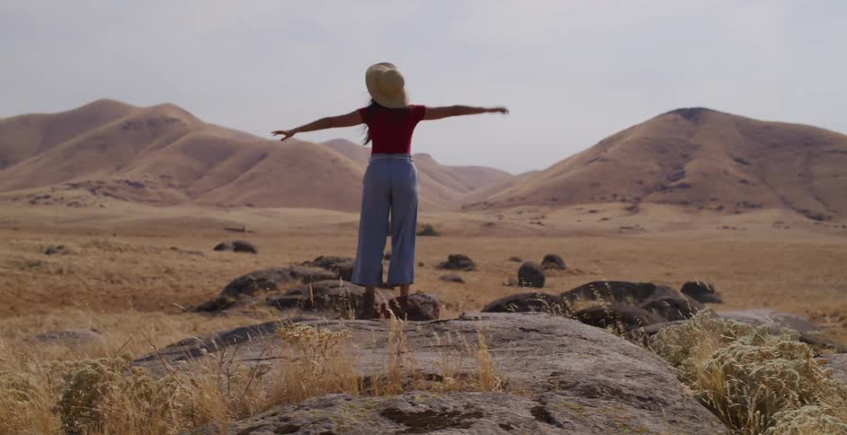 A free-spirited woman in a red shirt and hat joyfully dances and throws her hat in the air while standing on a boulder in a vast, arid valley