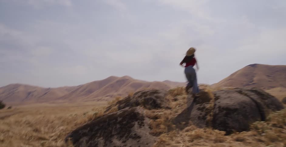 A young woman runs freely through a vast, dry, hilly landscape, reaching the top of a rock formation and opening her arms in triumph