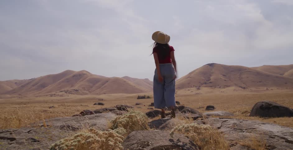 A woman in a hat runs up a rocky hill in a dry, desert landscape and triumphantly spreads her arms to the view