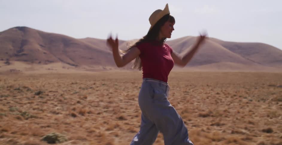 A carefree woman wearing a straw hat runs joyfully in slow motion through a vast, golden field with arid hills in the distance