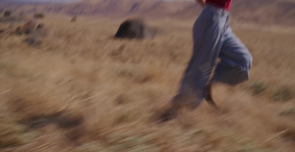A close-up tracking shot captures a woman's legs in wide pants running swiftly through a dry, golden field on a sunny day