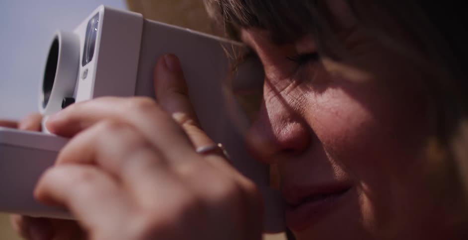 A woman's face is captured in an intimate slow-motion close-up as she takes a photo with an instant camera on a windy day