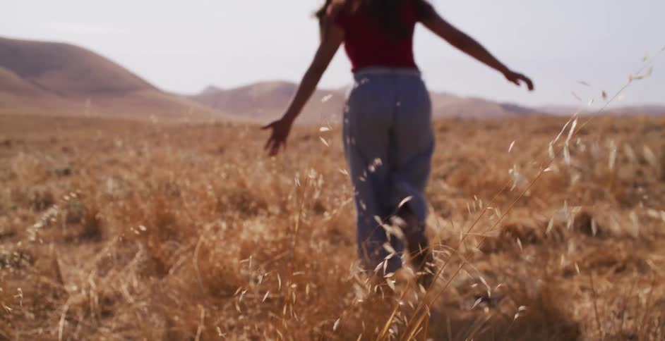 A woman in a straw hat runs away from the camera through a field of golden grass in slow motion