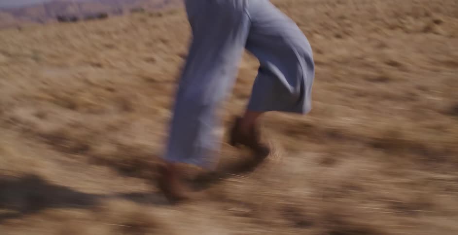 A joyful young woman in a straw hat runs and skips in slow motion through a dry, golden grassy field on a sunny day