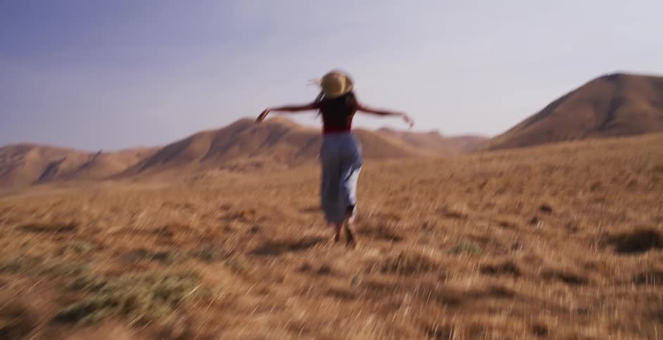 A carefree young woman in a sun hat runs with her arms outstretched through a vast, dry field toward rolling hills