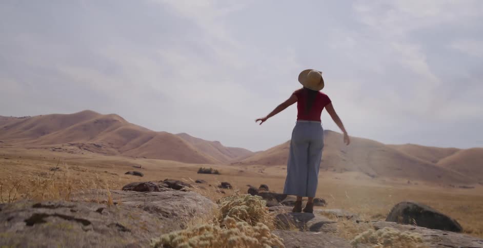 A young woman in a straw hat runs in slow motion up a rocky hill in a vast, golden landscape, then triumphantly spreads her arms to the sky