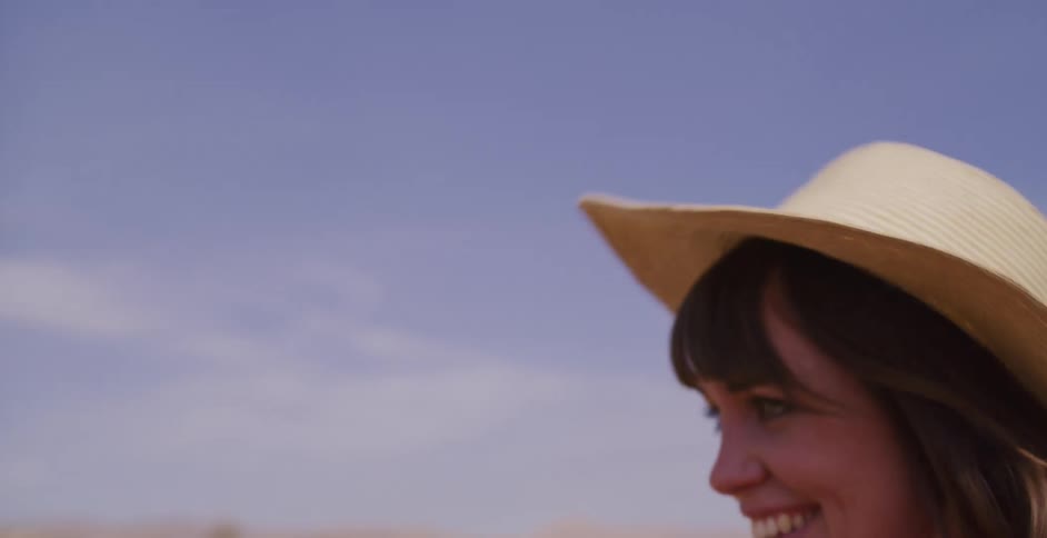 A carefree young woman in a straw hat smiles as she runs through a sunny, dry landscape in slow motion