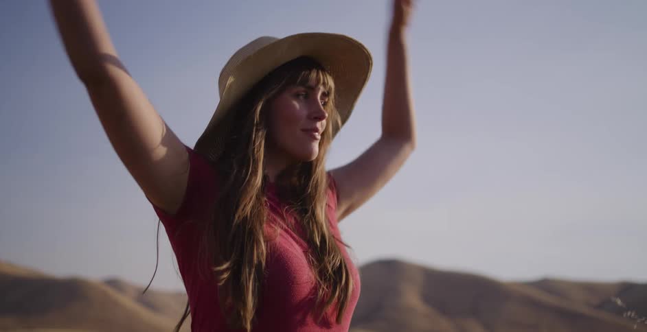 A serene young woman in a straw hat stretches her arms out, soaking in the warm golden hour sun in a tranquil desert landscape