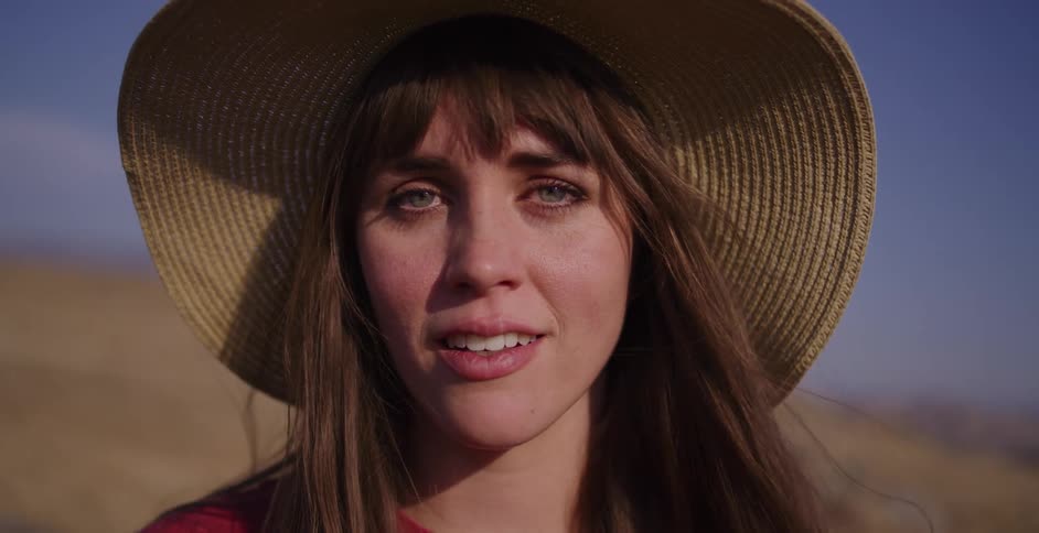 A serene young woman wearing a straw hat smiles with her eyes closed, then opens them and looks directly into the camera in slow motion