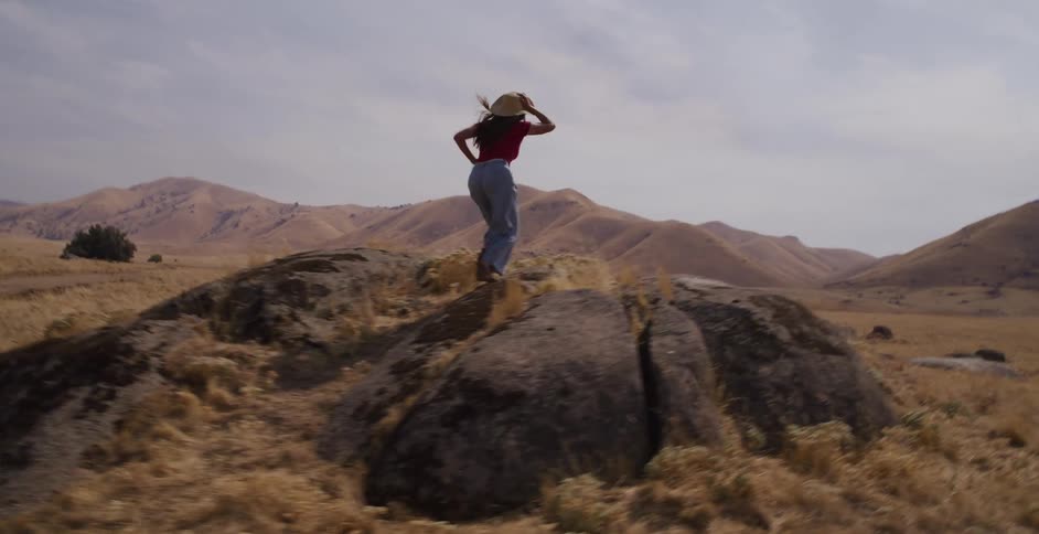 A woman runs freely through a dry, hilly landscape and stands on a boulder with her arms outstretched, enjoying the view