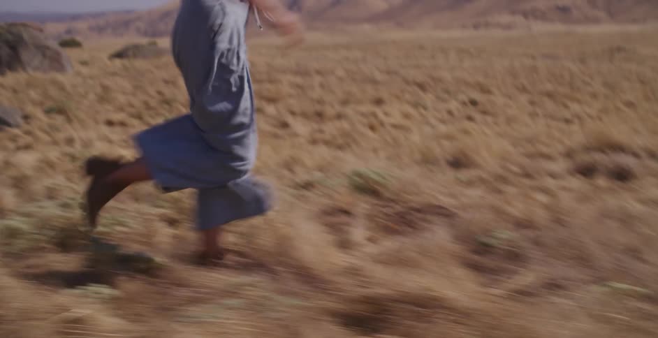 A happy young woman in a straw hat runs in slow motion through a golden field of tall grass with hills in the background