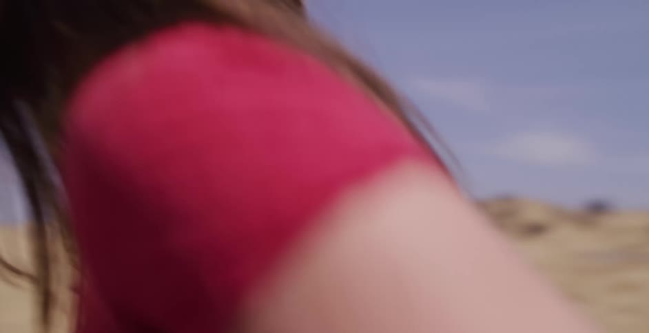 A smiling young woman in a straw hat holds it down while running through a sun-drenched, windy desert in slow motion