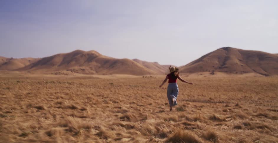 In slow motion, a free-spirited woman in a straw hat runs through a vast, golden field towards rolling hills on a sunny day