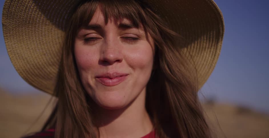 Slow motion close-up of a happy woman in a sun hat, smiling and closing her eyes to bask in the warm sunlight