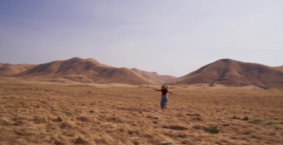 A carefree woman in a straw hat runs with her arms outstretched through a vast, dry golden field towards distant rolling hills