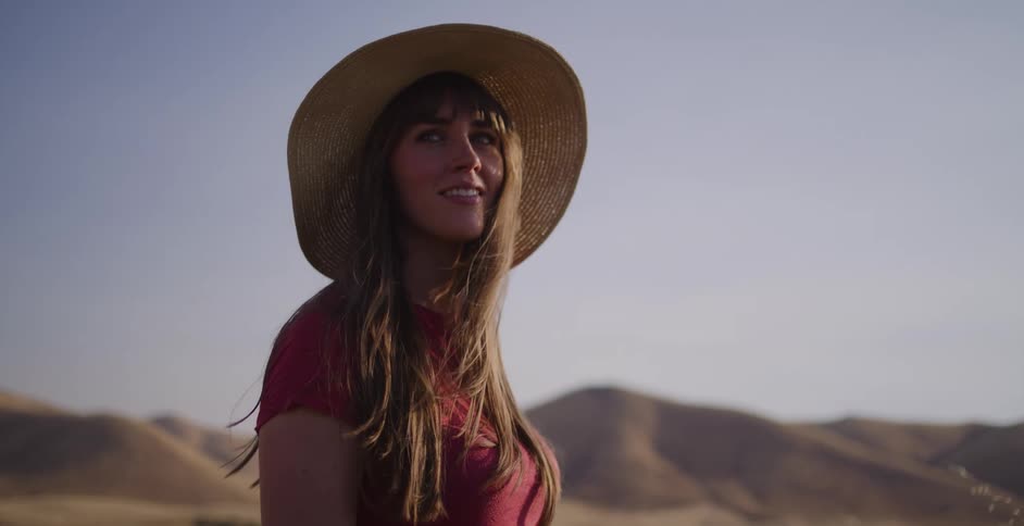 A contemplative woman wearing a sun hat enjoys the warm breeze in a golden field with rolling hills in the background